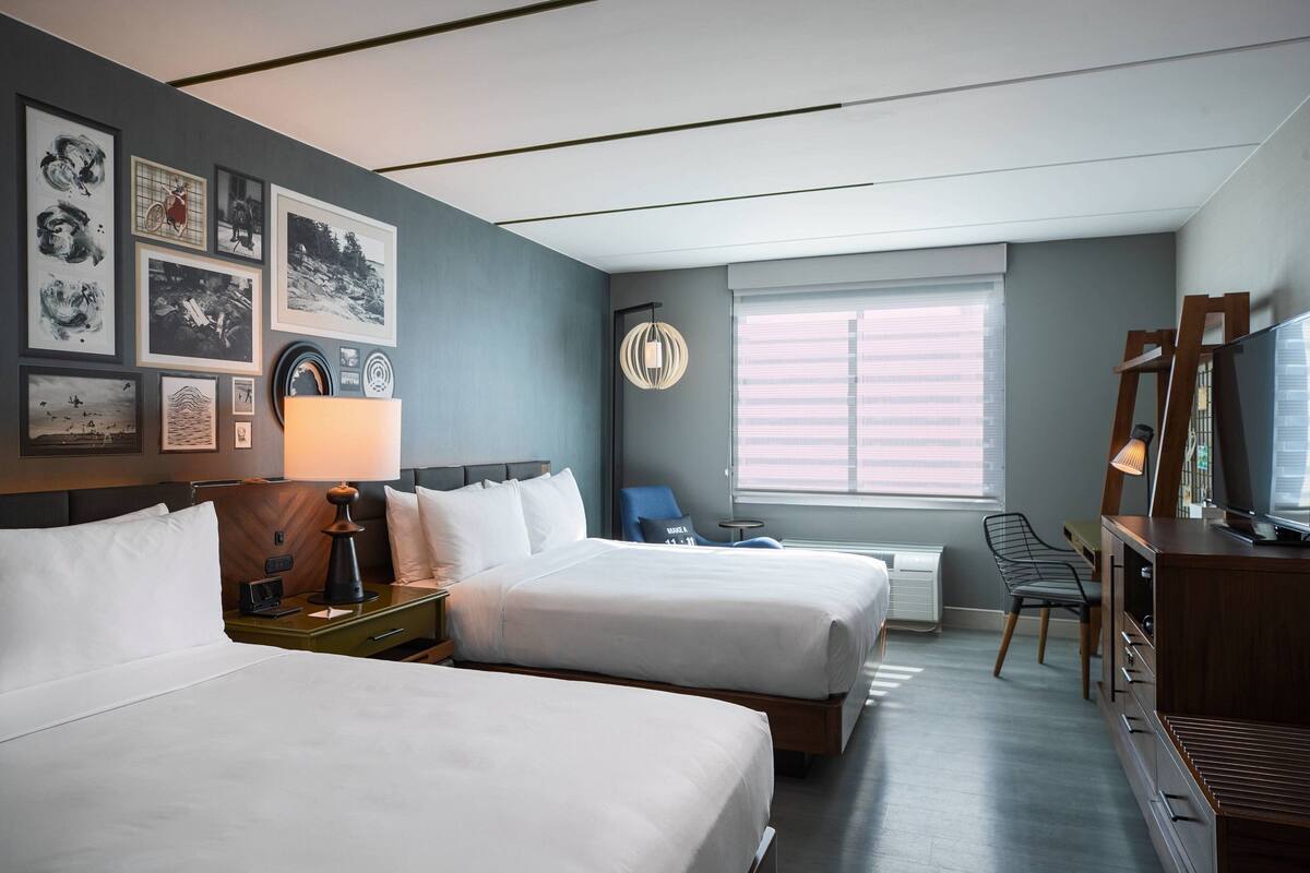 A hotel room with two beds, a desk, a chair, a TV, and a window with blinds.