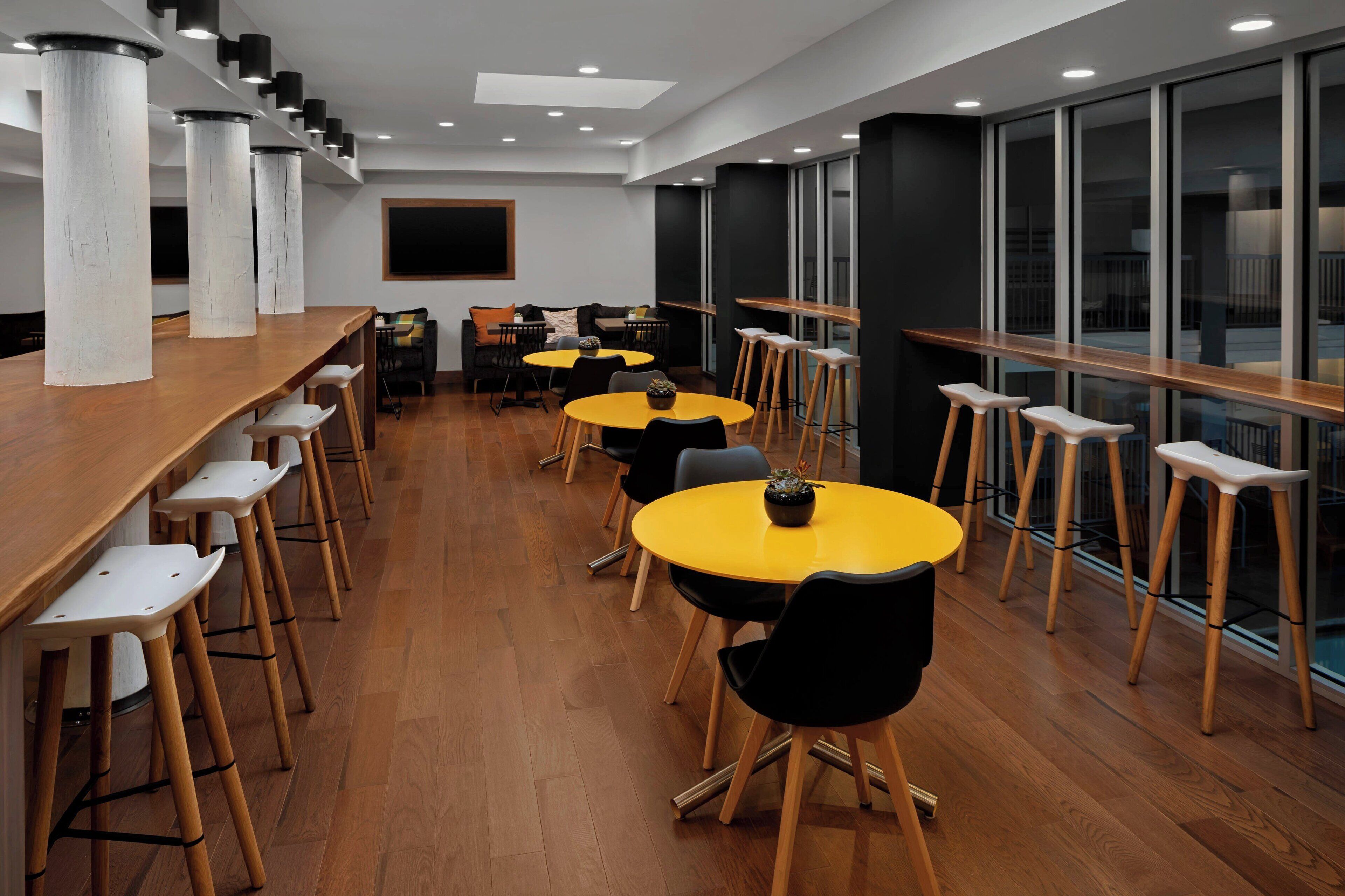A modern dining area with high wooden tables and chairs, large windows, and a clean, minimalist design.