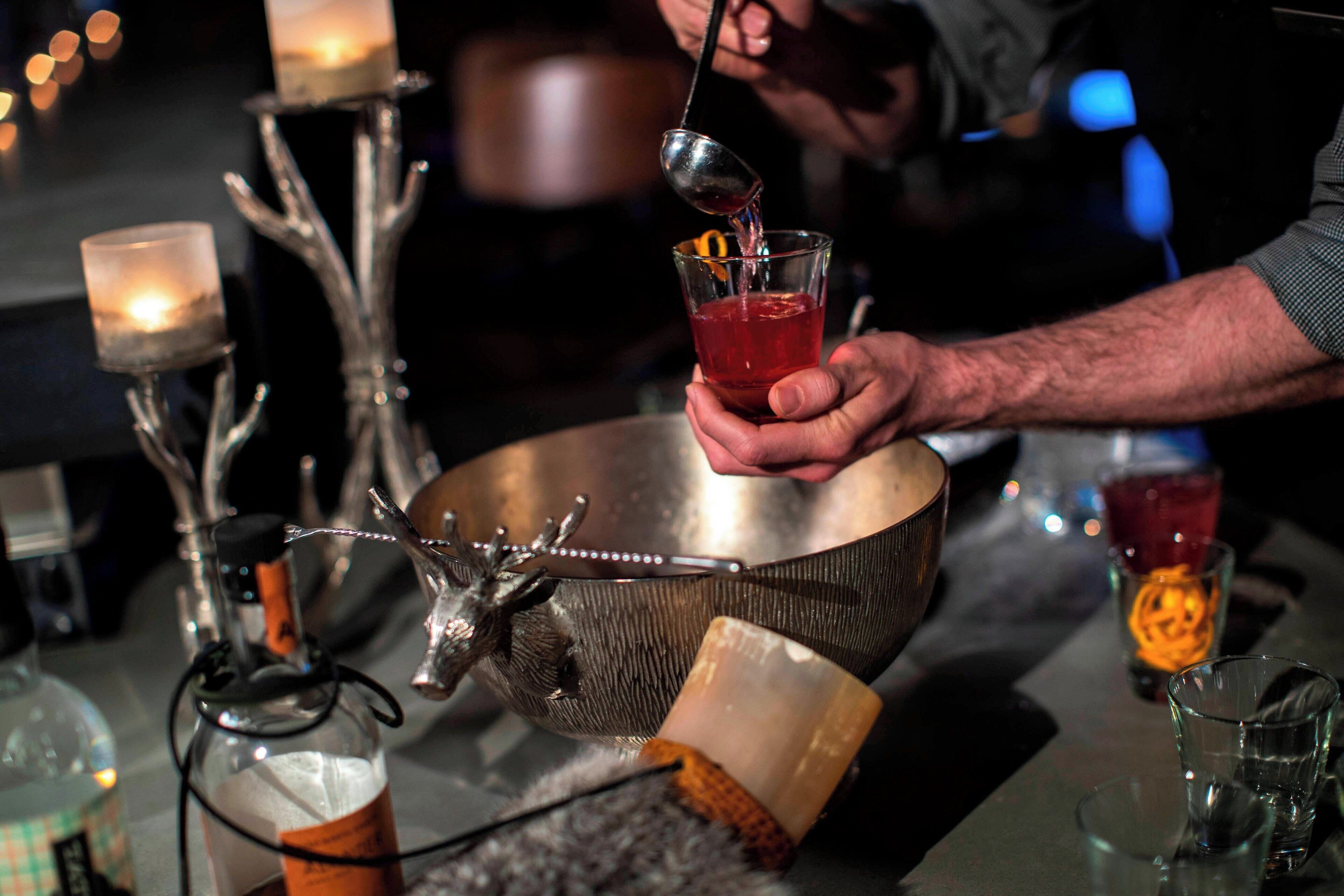 Un barman verse une boisson dans un verre, avec un bol en métal décoratif et une bougie en arrière-plan.