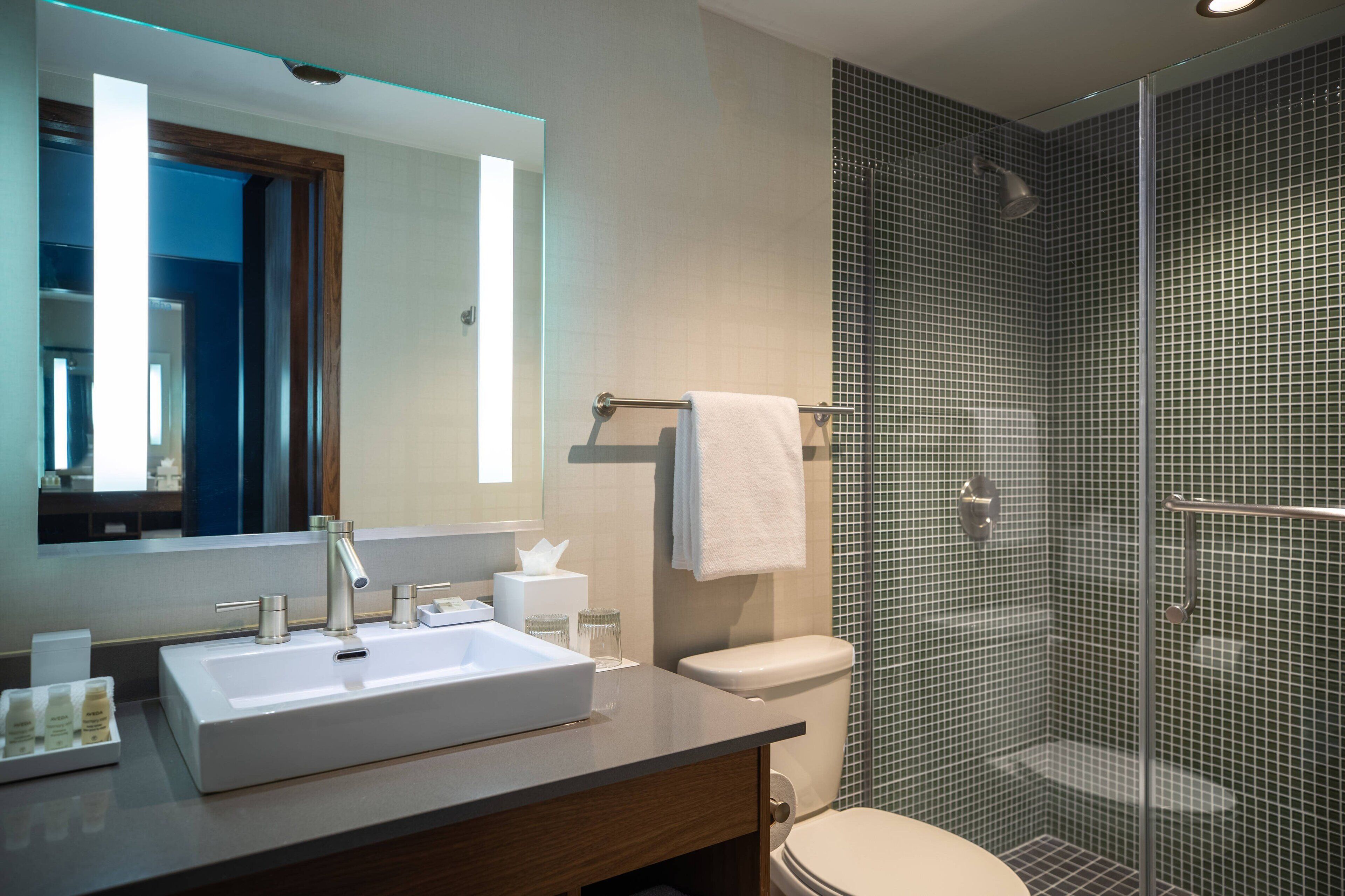 Un baño moderno con un espejo grande, un lavamanos blanco, un inodoro y una ducha con mampara de vidrio.