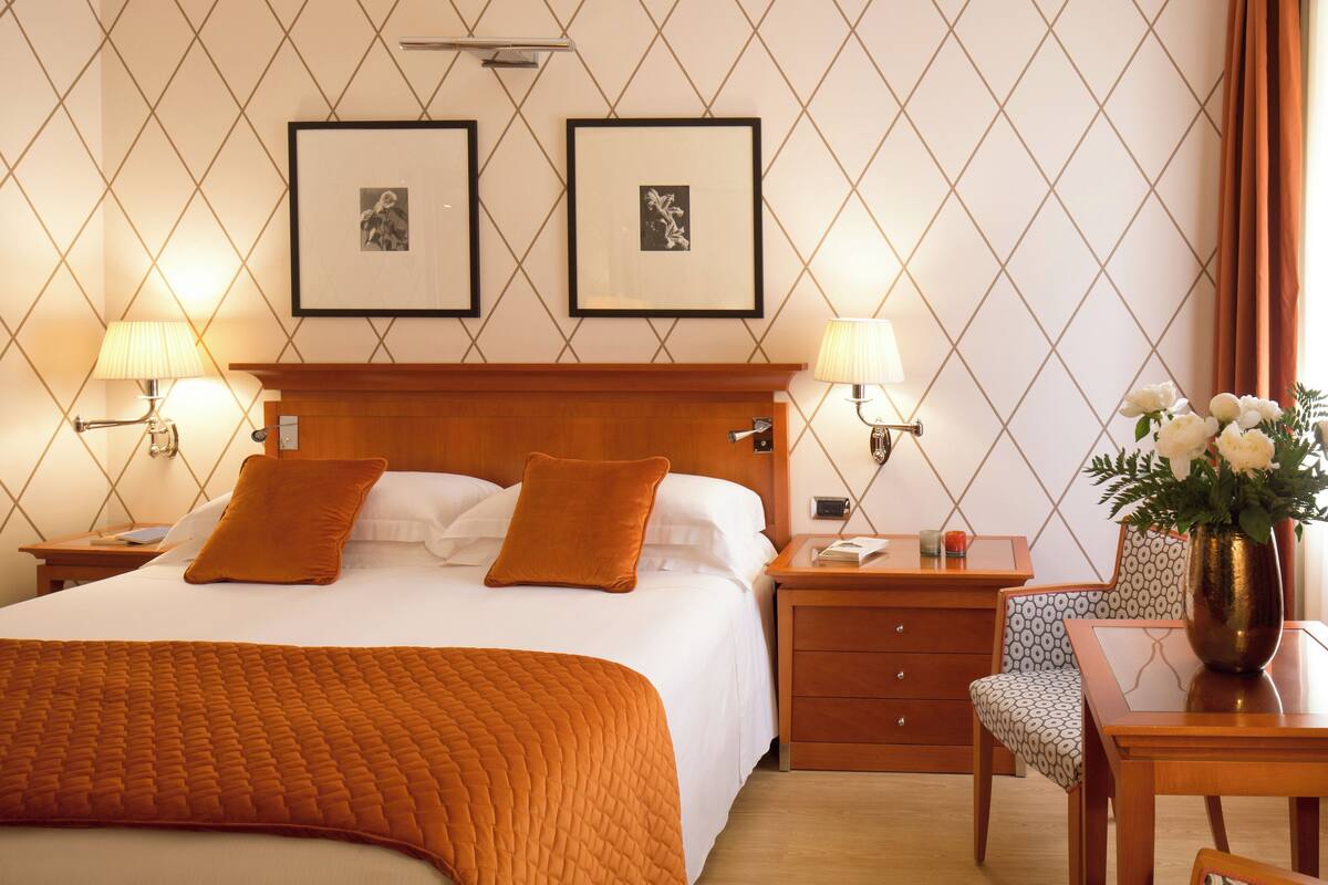 A hotel room with a large bed, two framed pictures on the wall, a nightstand with a lamp, a chair, and a table with a vase of flowers.