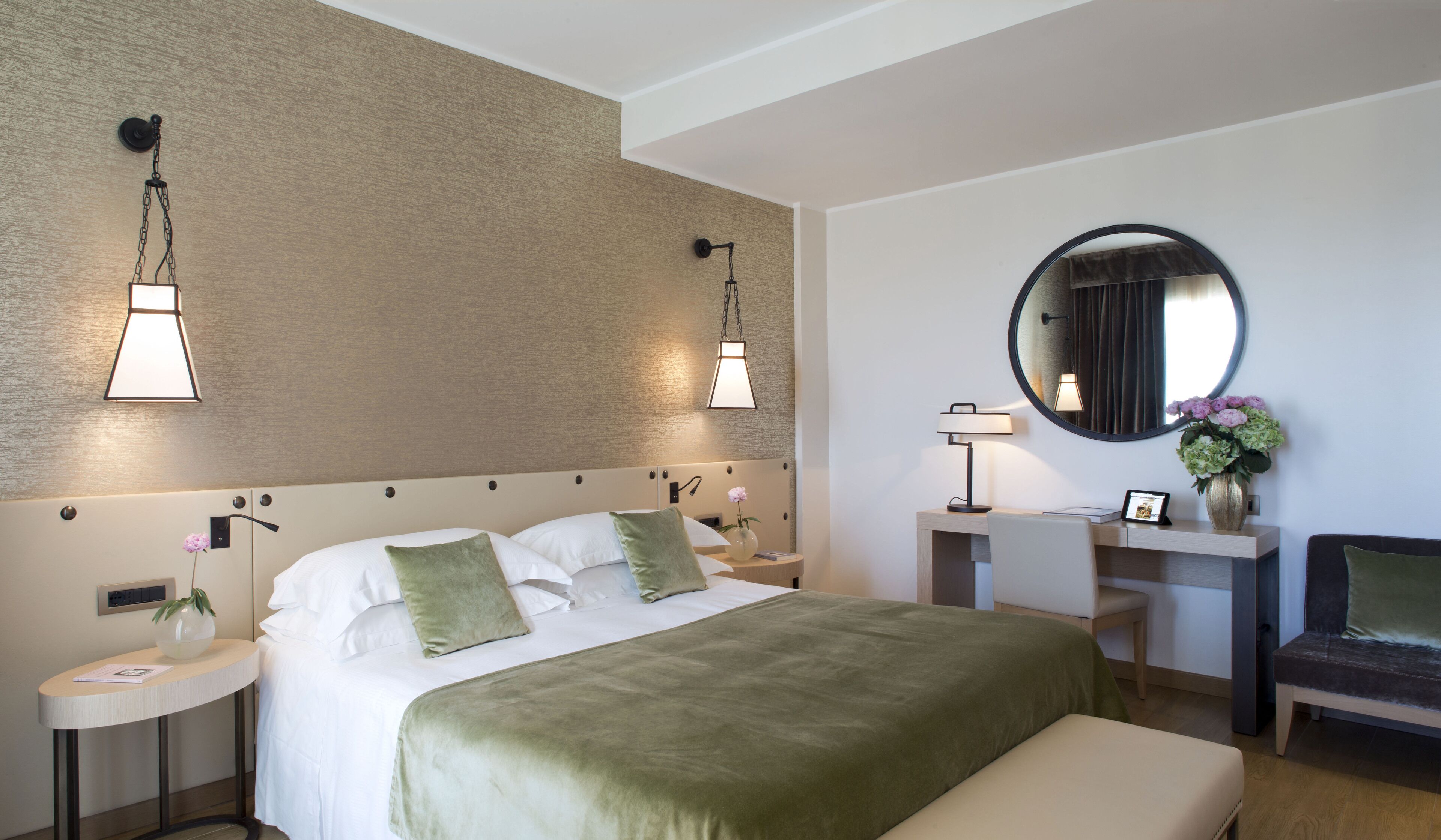 Une chambre d’hôtel avec un grand lit, des tables de chevet, un bureau, une chaise, un miroir rond et des lampes fixées au mur.