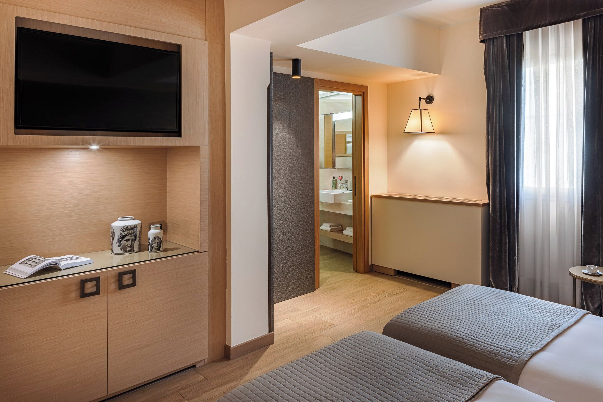 Habitación de hotel con una televisión de pantalla plana montada en la pared, una mesita de noche con lámpara y una cama con edredón gris.