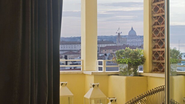 Un balcón con vista a un paisaje urbano, que incluye un edificio destacado con cúpula. Hay una silla de mimbre, una mesita con lámpara y una cama con vistas.