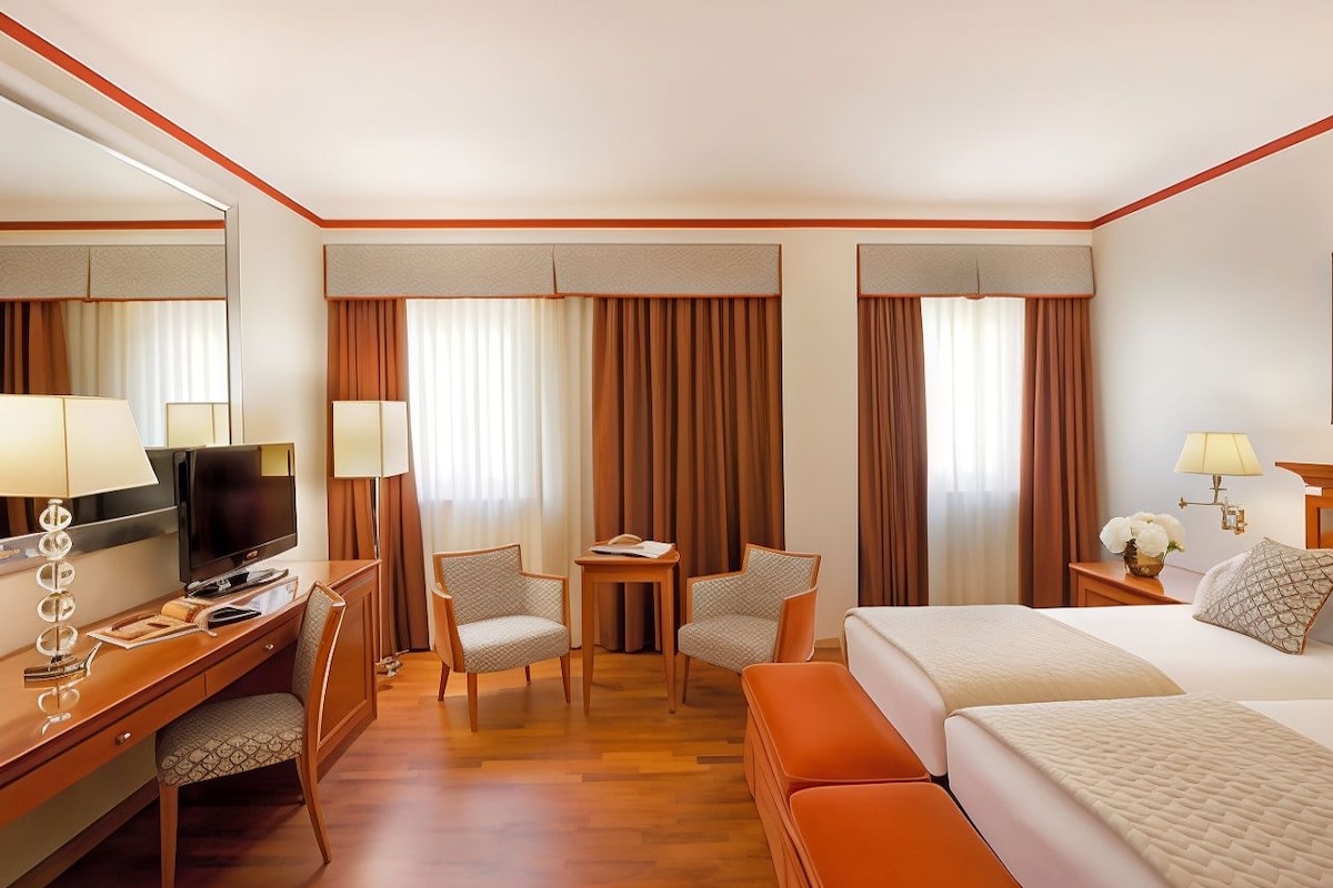 Une chambre d’hôtel avec un lit, un bureau avec une télévision, deux chaises, une petite table et des rideaux.