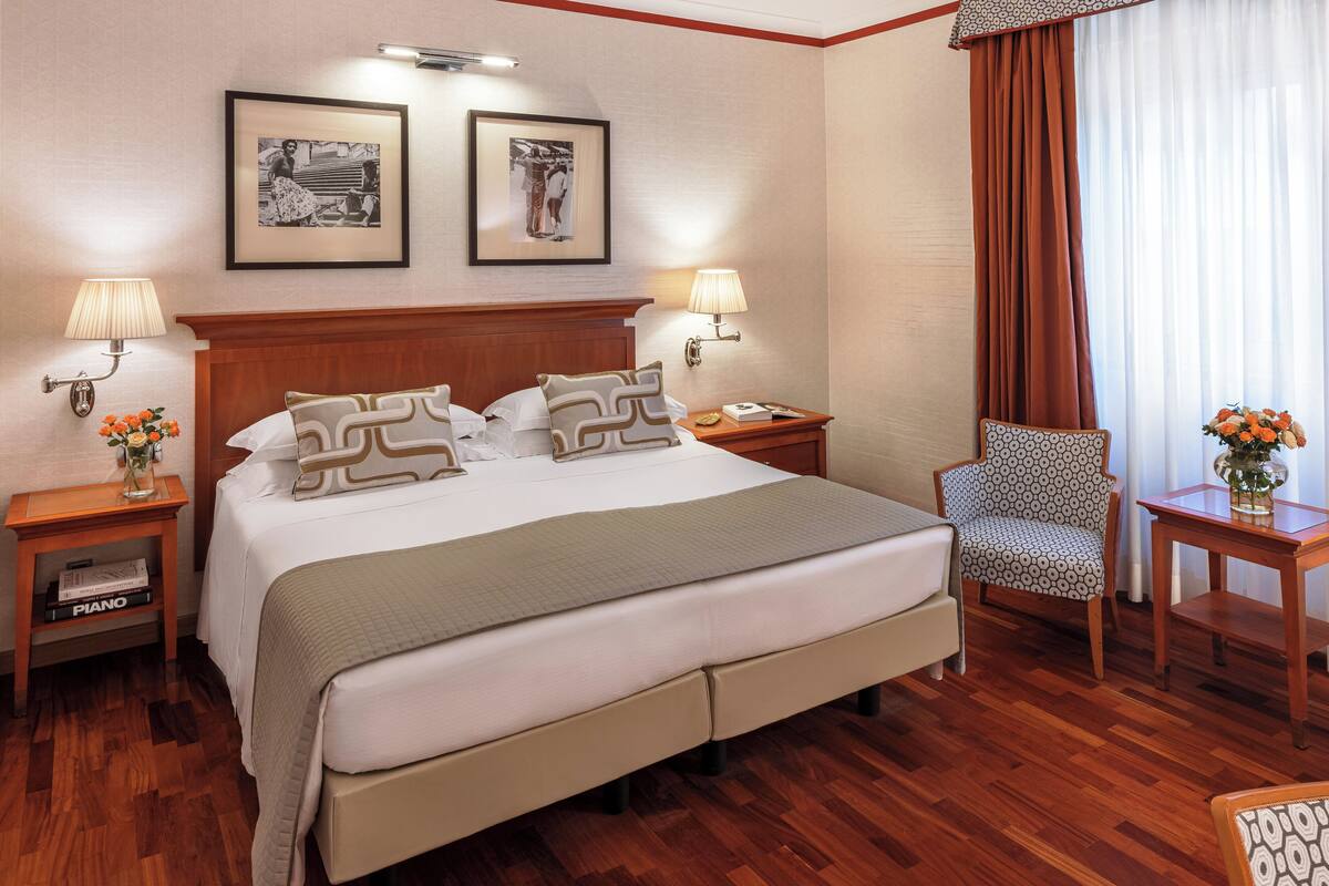 A hotel room with a large bed, two bedside tables with lamps, a chair, a small table with a vase of flowers, and two framed pictures on the wall.
