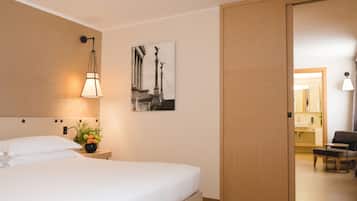 A hotel room with a bed, a nightstand with a lamp and a vase of flowers, a wall-mounted picture, and a wooden door leading to another room.