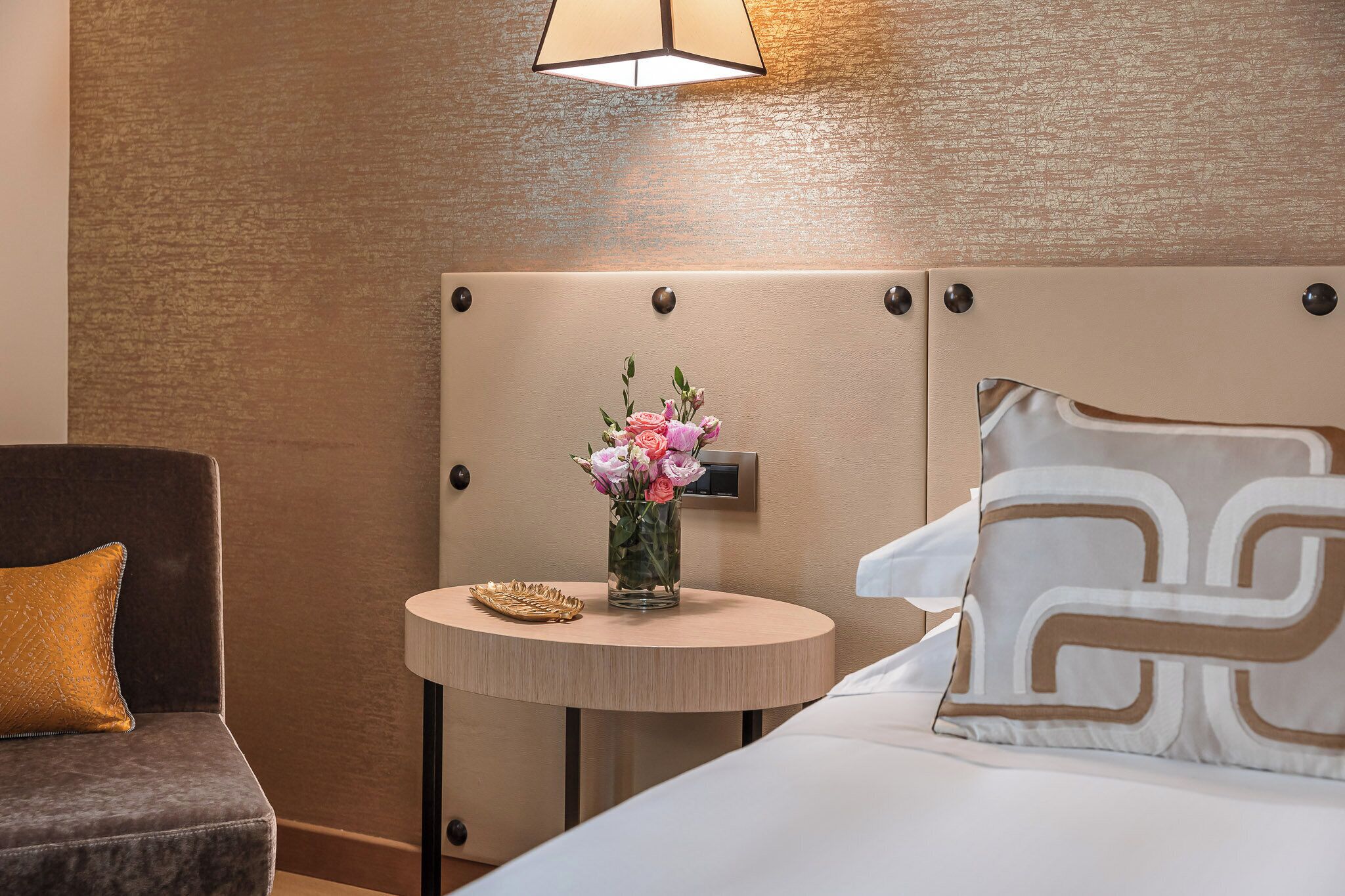 A hotel room with a bed, a bedside table with a vase of flowers, a lamp, and a chair.