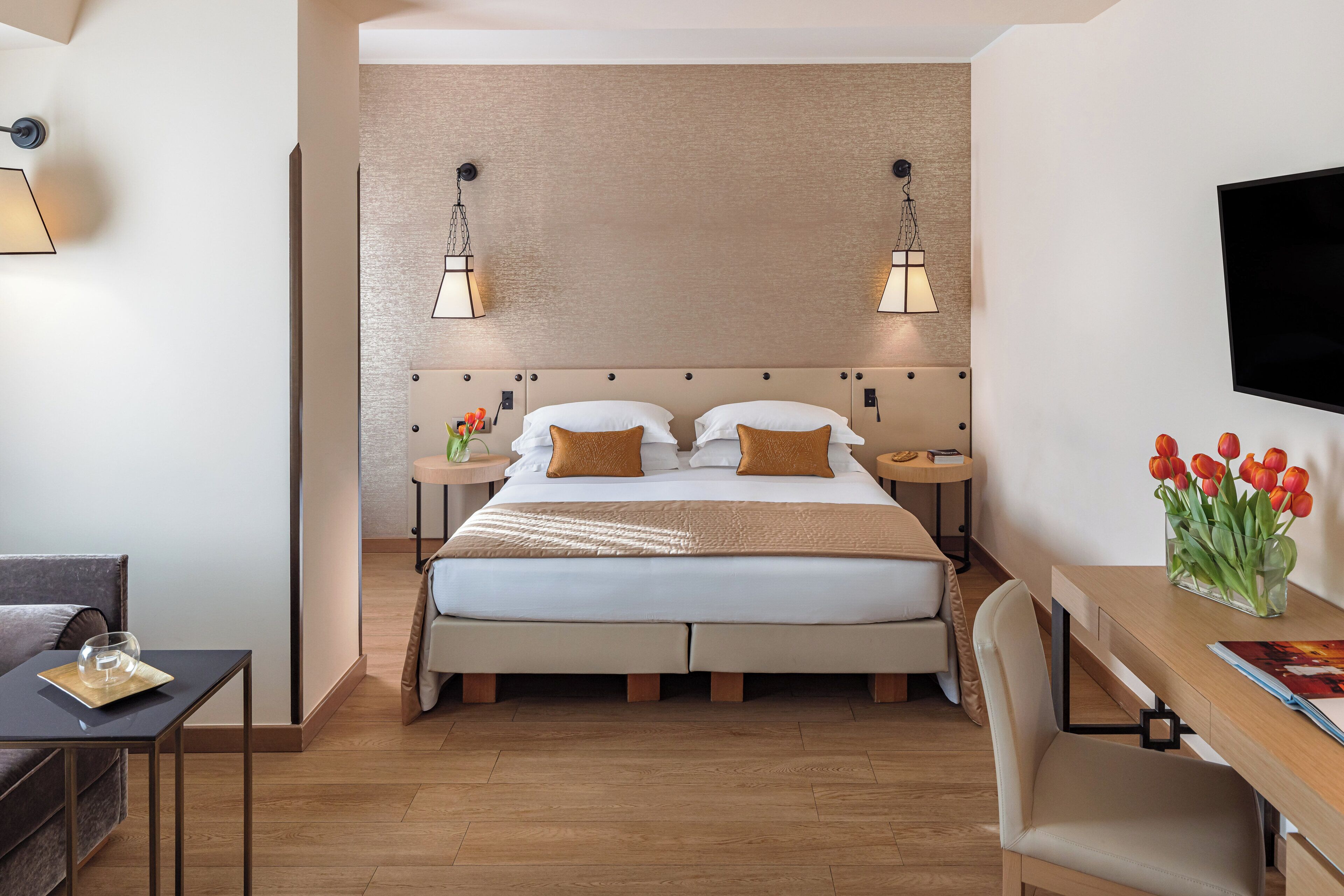 A modern hotel room with a large bed, a desk, a chair, a TV, and a vase of tulips.