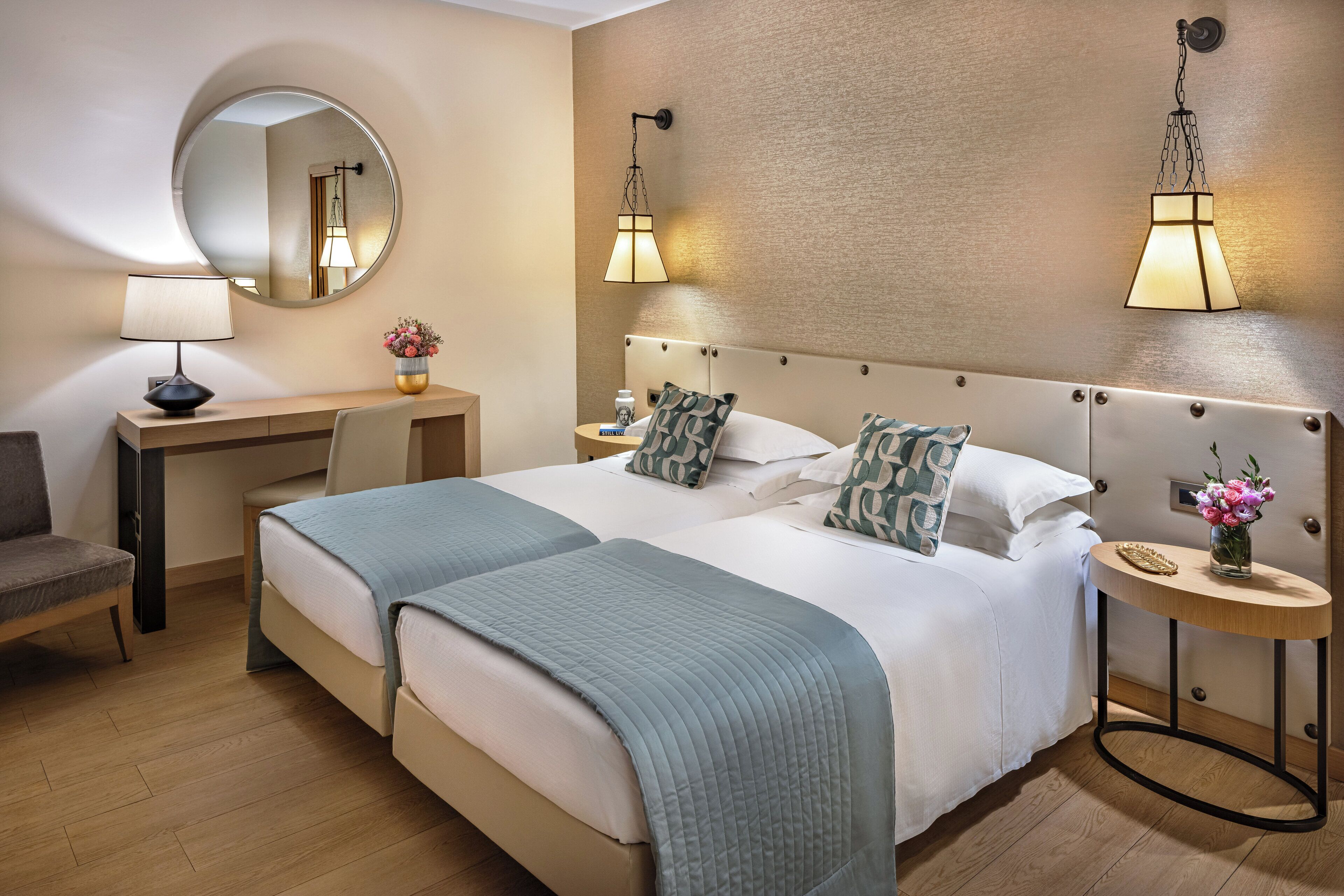 A hotel room with two beds, a desk, a chair, a round mirror, and two wall-mounted lamps.