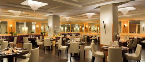A spacious hotel dining area with multiple round tables and chairs, large chandeliers, and a bar area.