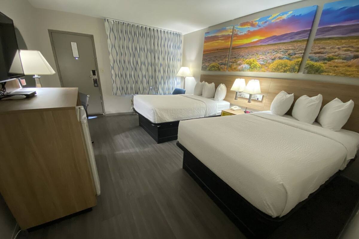 A hotel room with two beds, a desk, a TV, and a painting of a sunset.