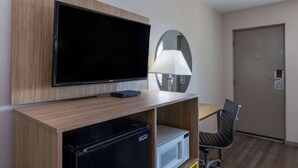 A hotel room with a flat-screen TV mounted on a wooden shelf, a microwave, a mini-fridge, a chair, a lamp, and a door. - Days Inn by Wyndham Roswell (Roswell)