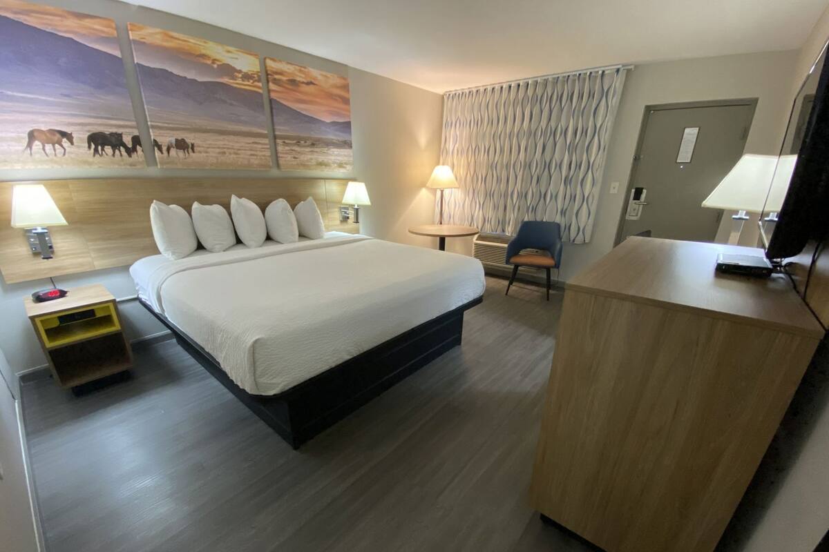 A hotel room with a large bed, two bedside lamps, a desk with a computer, a chair, and a wall with a sunset painting.