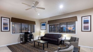 A living room with a brown leather sofa, a gray armchair, a coffee table, and a TV on a stand. There are two windows with blinds, a ceiling fan, and a rug on the floor. - Days Inn by Wyndham Roswell (Roswell)