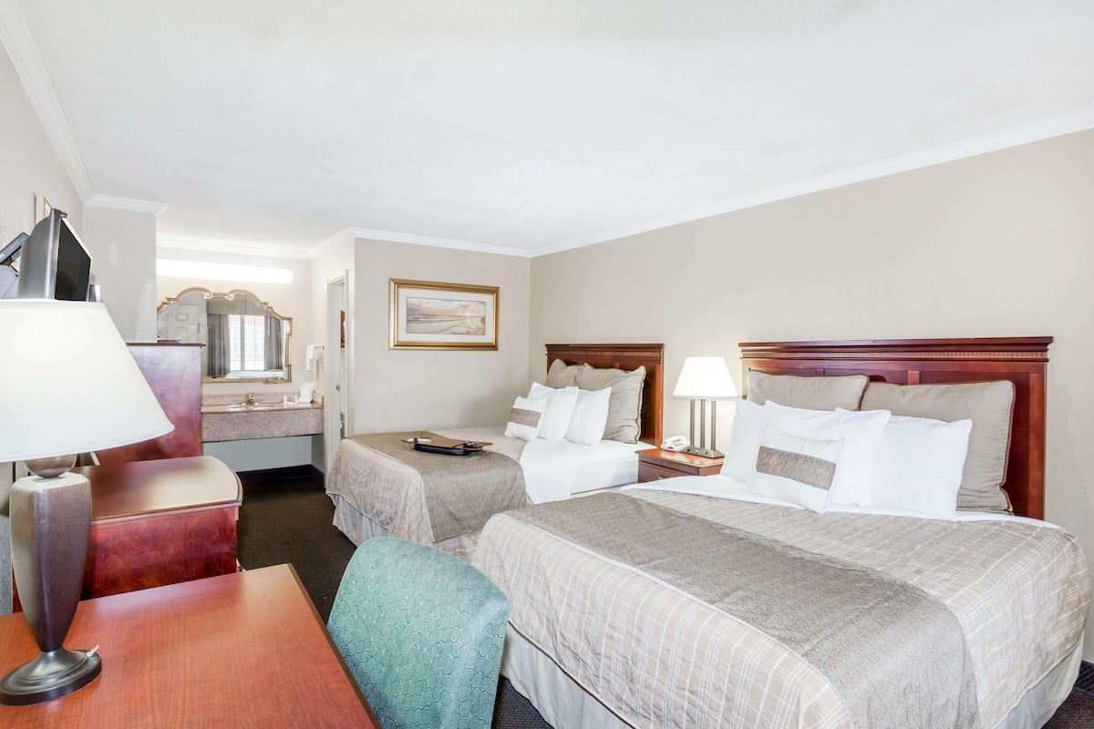 A hotel room with two beds, a desk, a chair, a lamp, a mirror, and a painting on the wall.