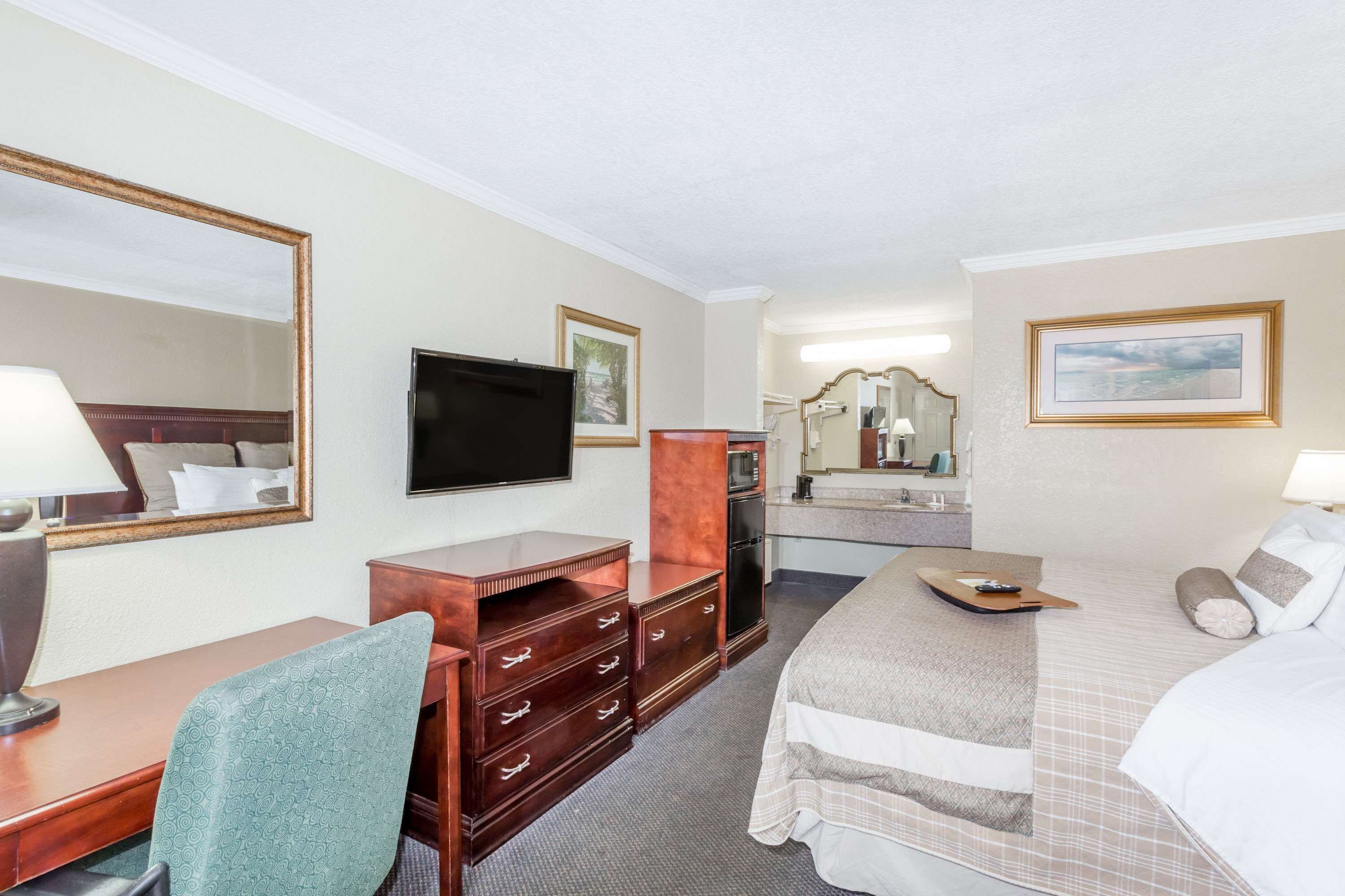 A hotel room with a bed, a desk with a chair, a television, a dresser, a mirror, and a bathroom with a sink and mirror.
