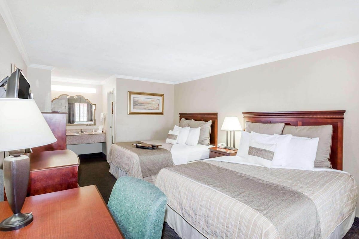 A hotel room with two beds, a desk, a chair, a lamp, and a painting on the wall.