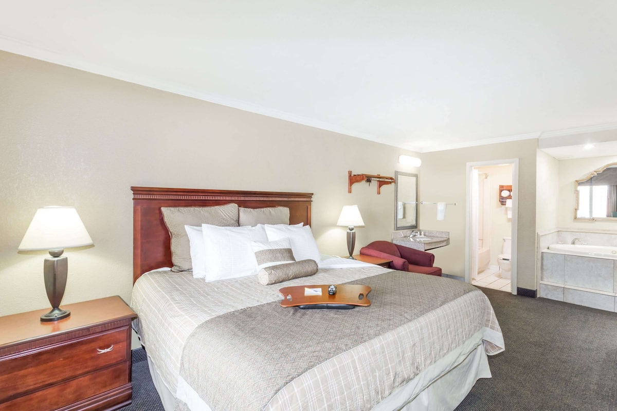 A hotel room with a large bed, two bedside tables, a desk, a chair, a bathroom with a sink and mirror, and a door leading to another room.