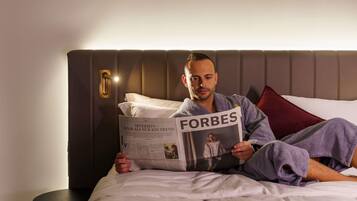 Una persona leyendo el periódico Forbes en una habitación de hotel con una cama moderna, una mesita de noche y una lámpara de pared.