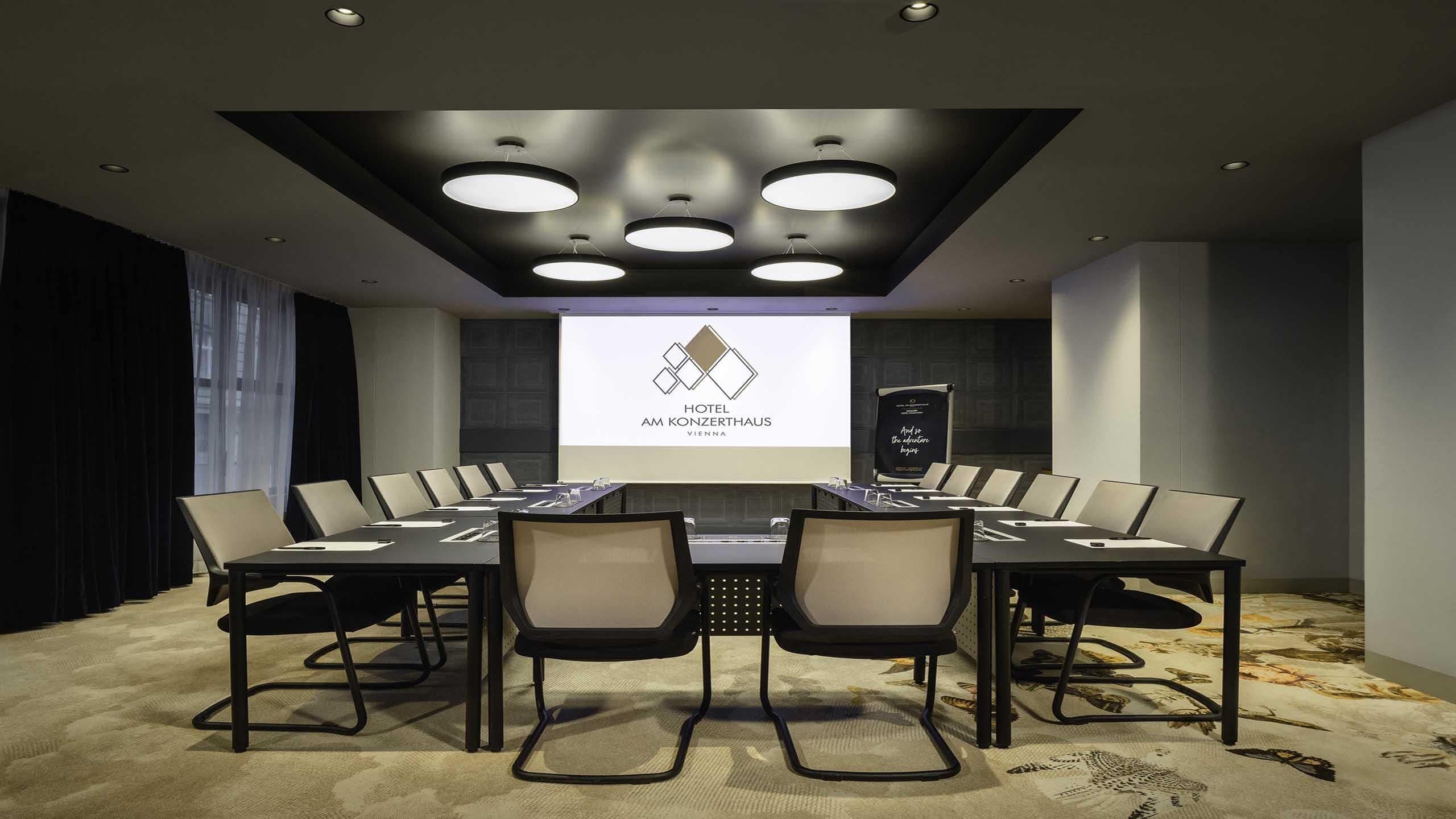A conference room with a large table, chairs, a projector screen, and a logo for Hotel Am Konzerthaus Vienna.