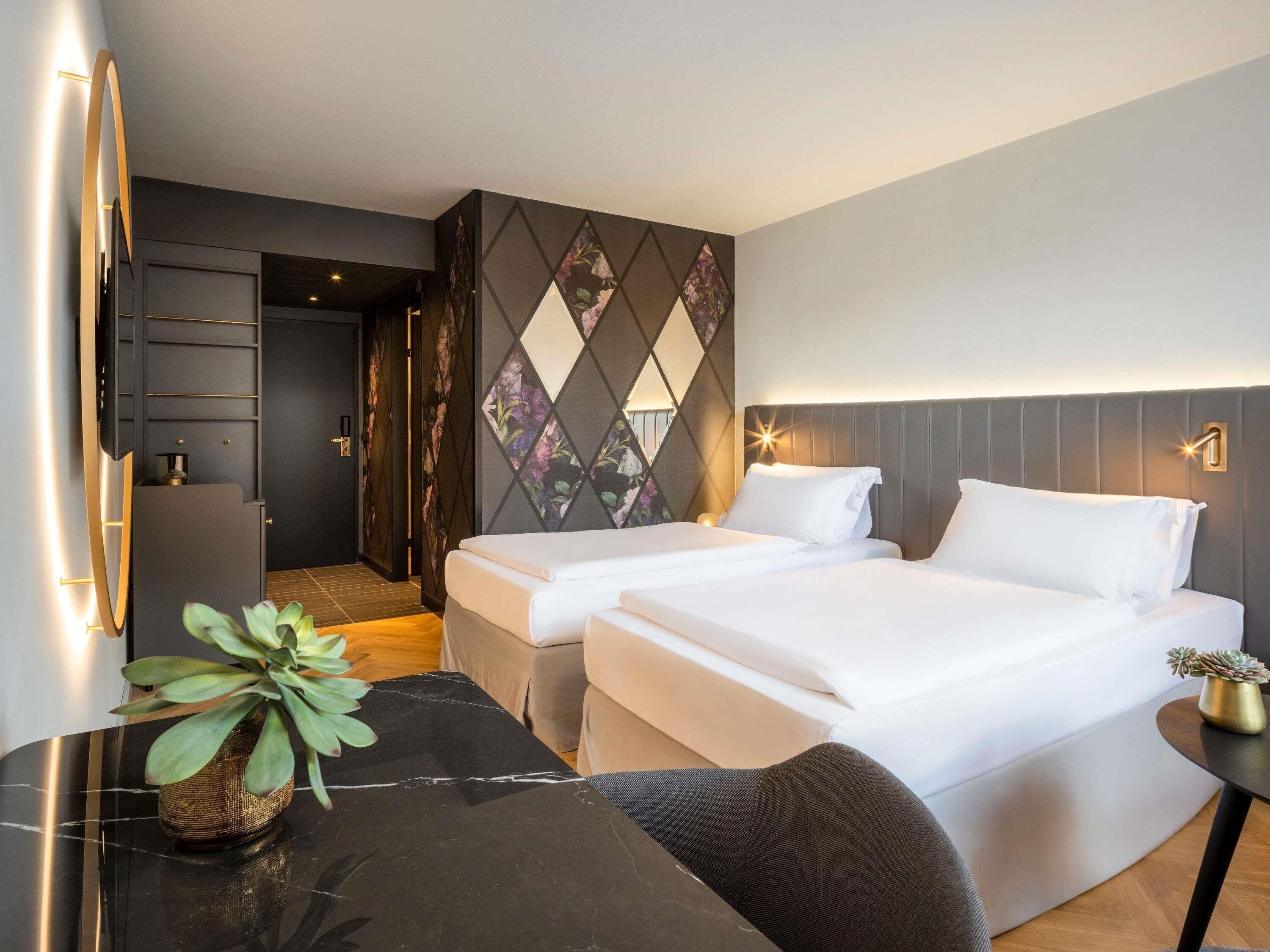 Quarto de hotel moderno com duas camas, uma mesinha e um vaso de planta.