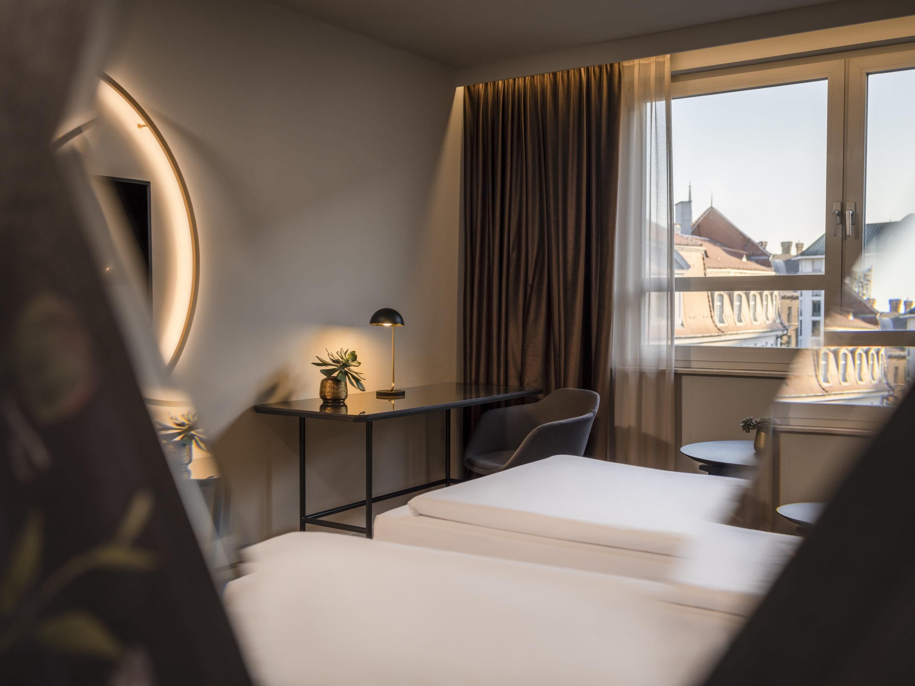Et moderne hotelværelse med seng, skrivebord, stol, lille bord og et vindue med gardiner.