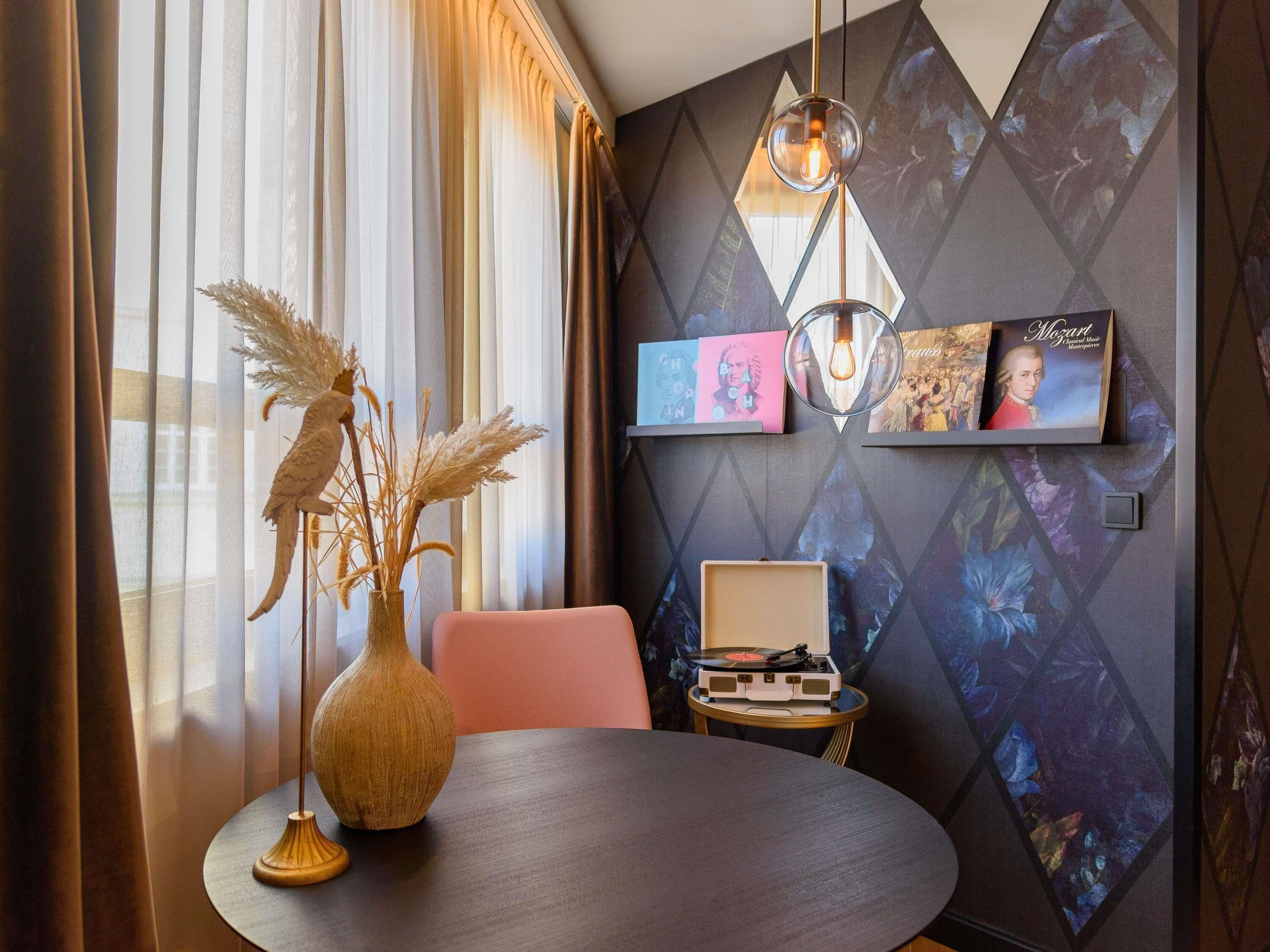 A modern room with a round table, a vase with dried plants, a small side table with a record player, and a wall with geometric patterns and framed pictures.