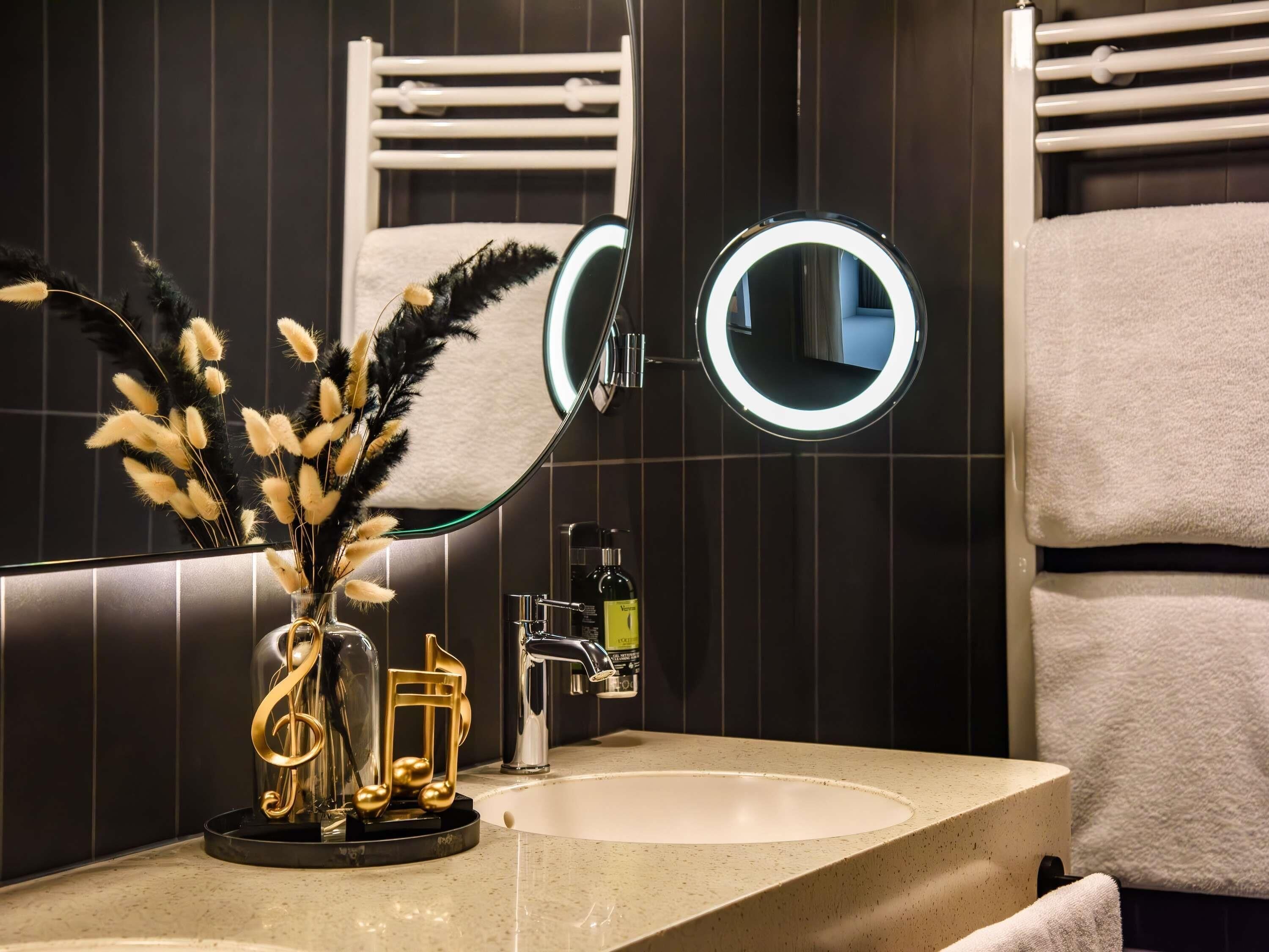Een badkamer met een wastafel, een spiegel met verlichting, een handdoekhouder en een decoratieve vaas met gouden noten.