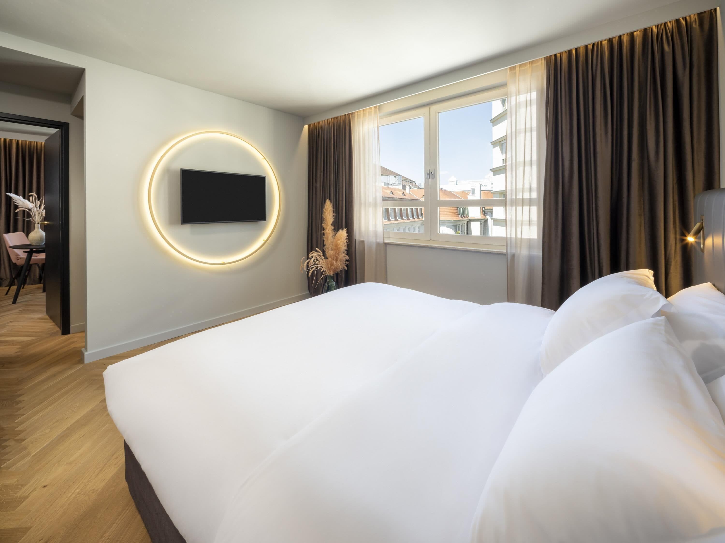 Et moderne hotelværelse med en stor seng, et rundt, vægmonteret TV, udsigt til bygninger gennem vinduet og trægulv.