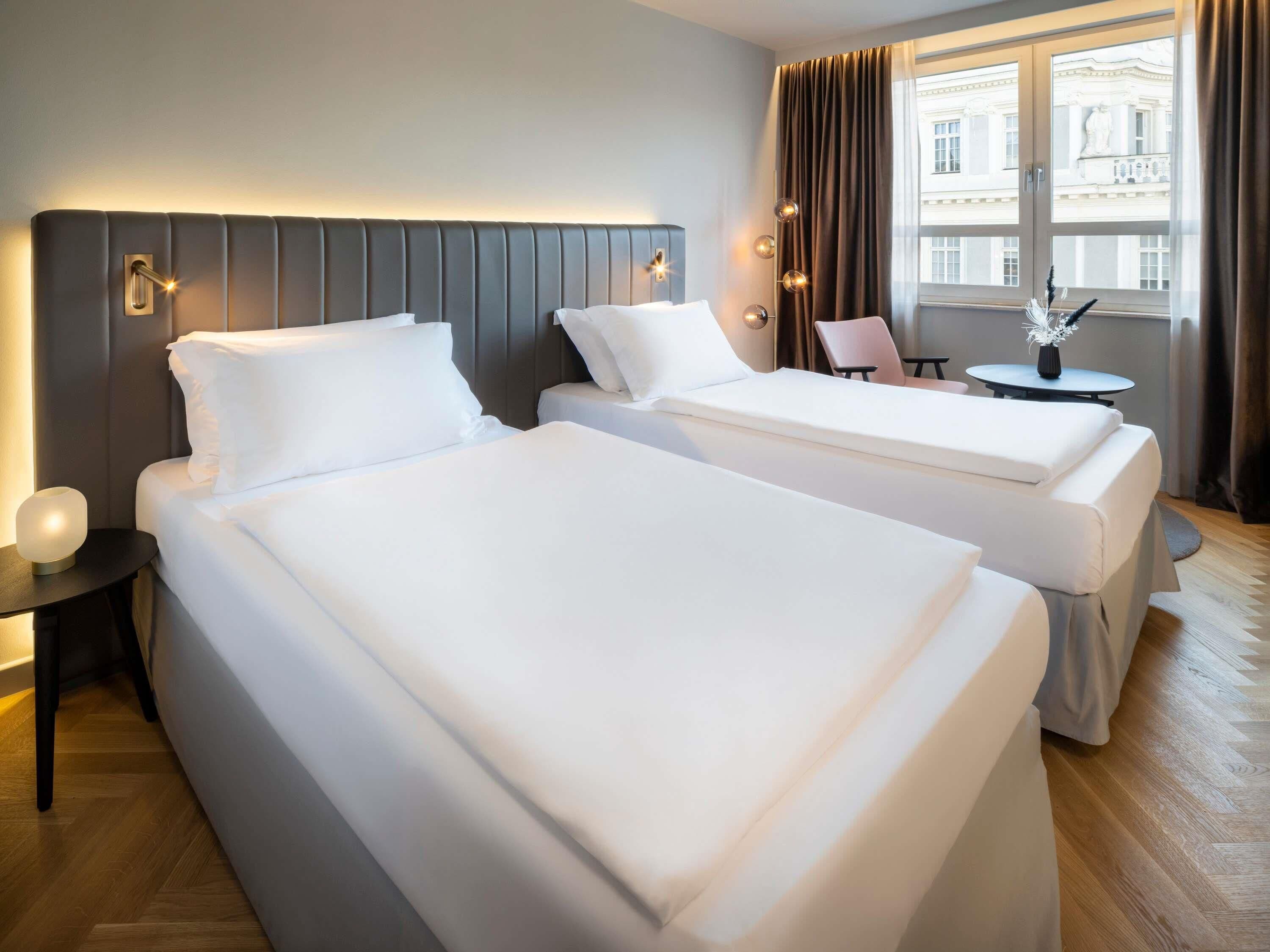 Habitación de hotel con dos camas, una mesita con un florero y vista a un edificio a través de amplias ventanas.