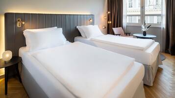 Habitación de hotel con dos camas, una mesita con un florero y vista a un edificio a través de amplias ventanas.