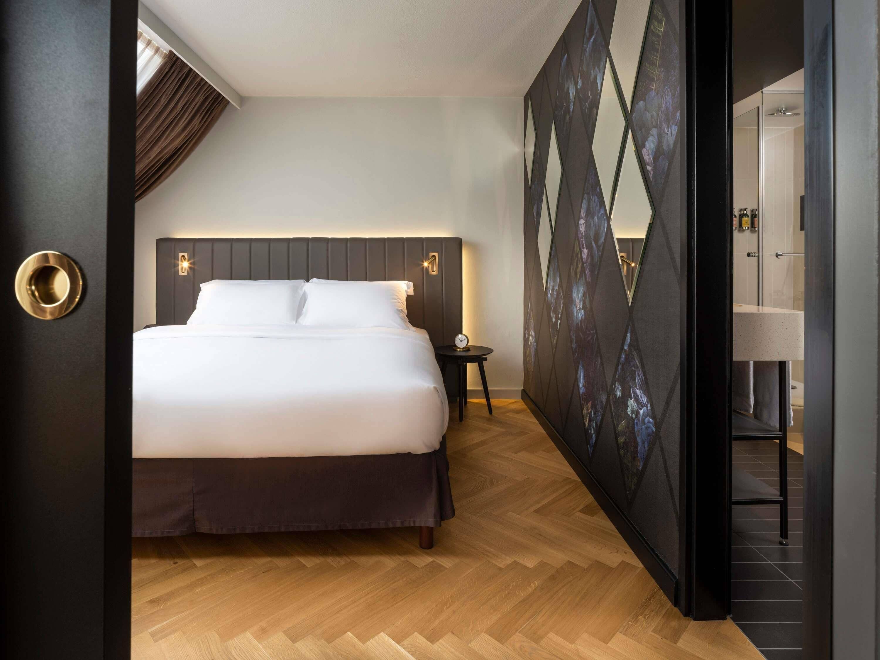 A modern hotel room with a large bed, a nightstand, a wall-mounted clock, and a geometric-patterned wall.