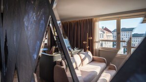A modern living room with a sofa, a coffee table, and a lamp. There is a window with a view of buildings outside. - Hotel Am Konzerthaus - MGallery Collection (Vienna)