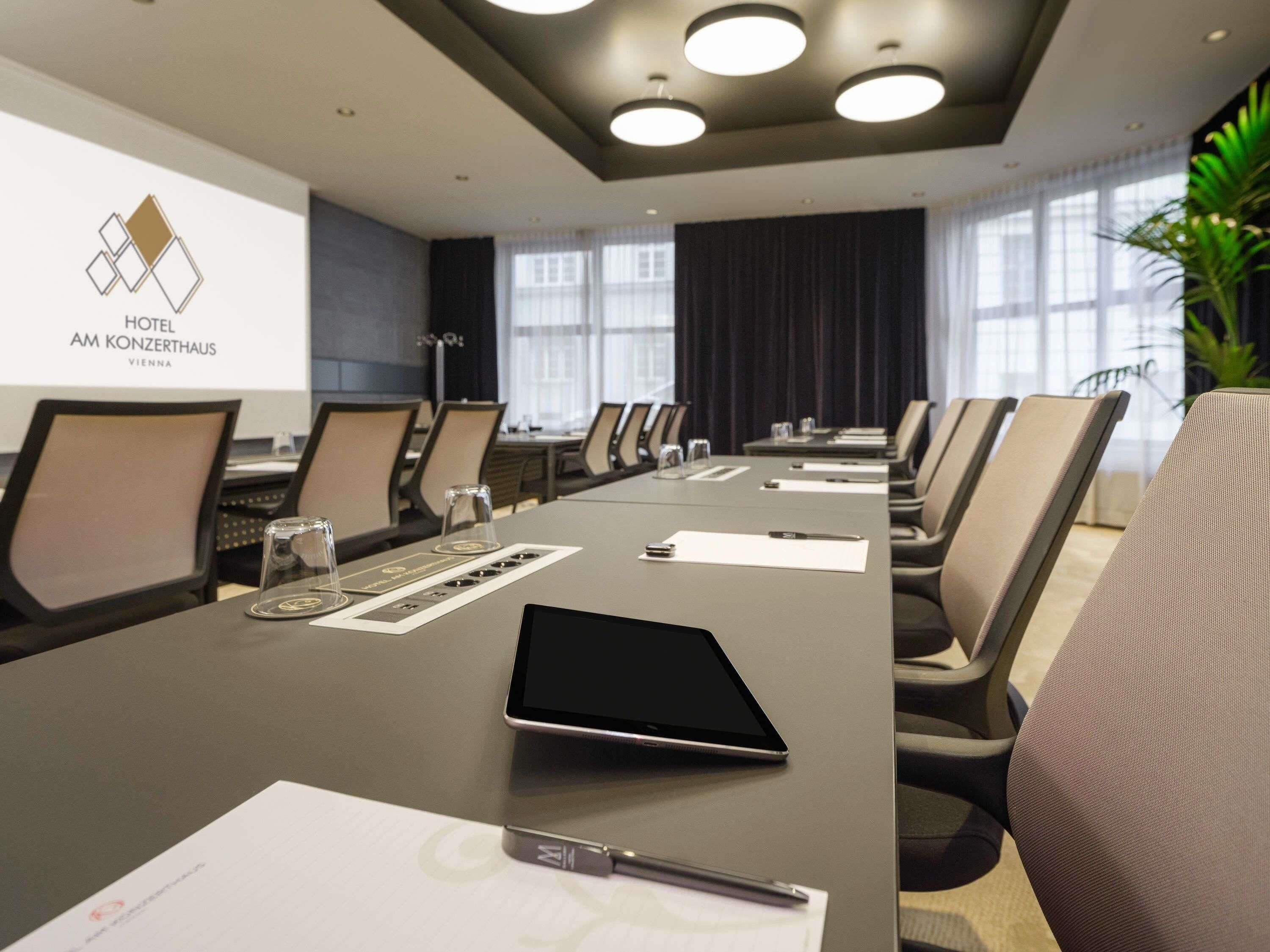 Una sala de conferencias con una larga mesa, sillas, una tableta y una pantalla de proyector que muestra el nombre del hotel.