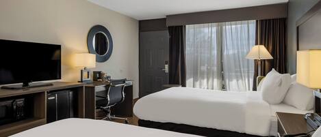 Habitación de hotel con dos camas, televisor, escritorio con lámpara y vistas al exterior a través de amplias ventanas.