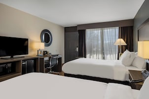 Habitación de hotel con dos camas, televisor, escritorio con lámpara y vistas al exterior a través de amplias ventanas.