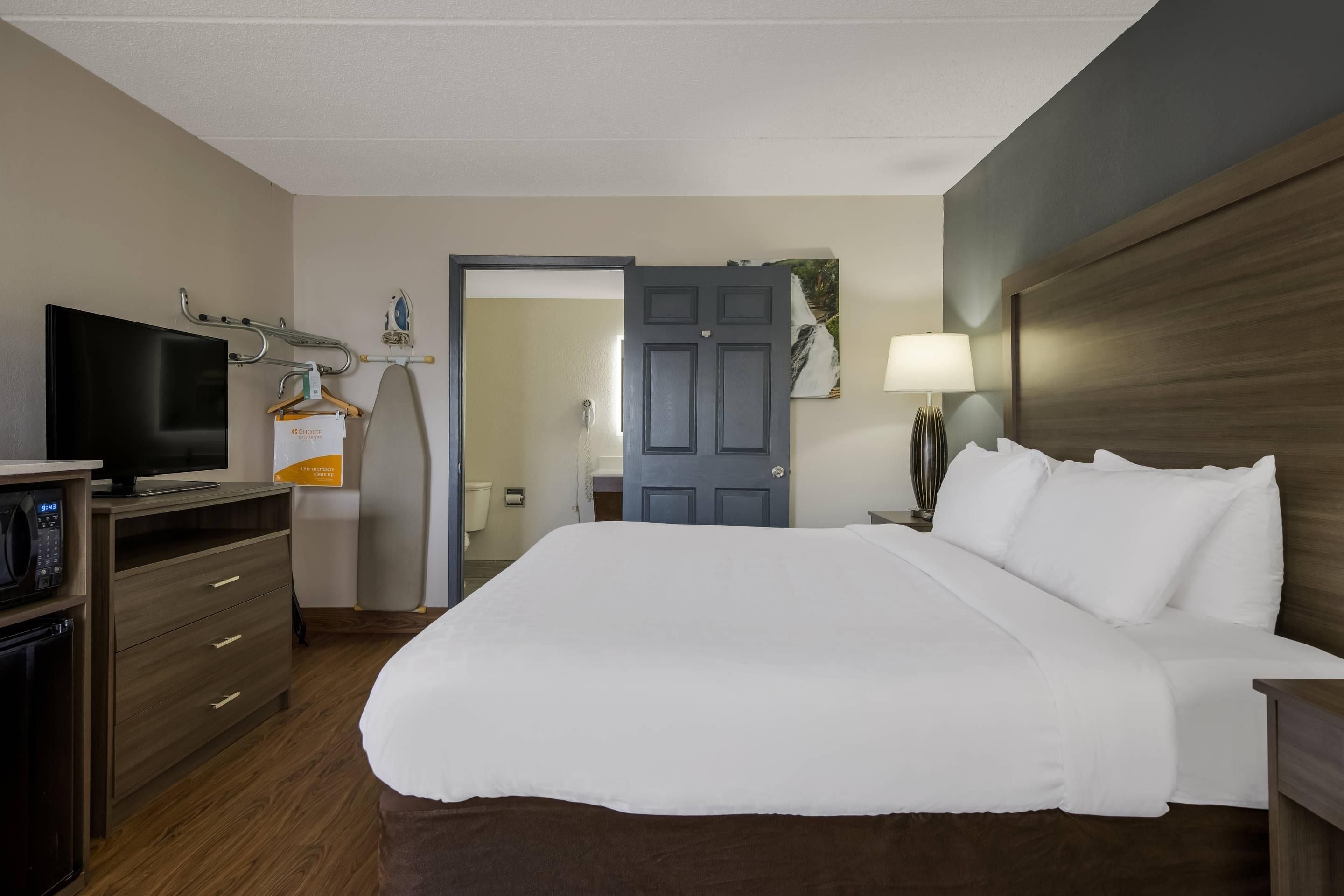 A hotel room with a bed, a TV, a desk with a computer, a lamp, and a door leading to a bathroom.