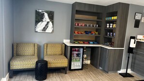 A seating area with patterned chairs, a small trash bin, a refrigerator stocked with beverages, snack shelves, and a wall-mounted picture of a waterfall.