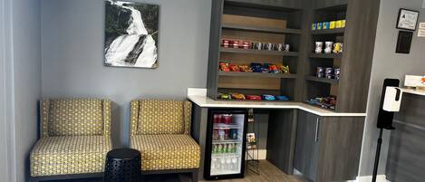 A seating area with patterned chairs, a small trash bin, a refrigerator stocked with beverages, snack shelves, and a wall-mounted picture of a waterfall.