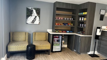 Un área de estar con sillas estampadas, un pequeño cubo de basura, un refrigerador lleno de bebidas, estantes con aperitivos y un cuadro colgado en la pared de una cascada.
