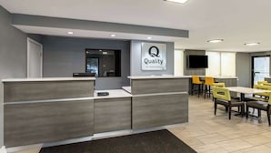 Zona de recepción de hotel con diseño moderno, que incluye un mostrador de recepción, un letrero que dice “Quality by Choice Hotels” y un área de comedor con sillas amarillas y un televisor de pantalla plana montado en la pared.