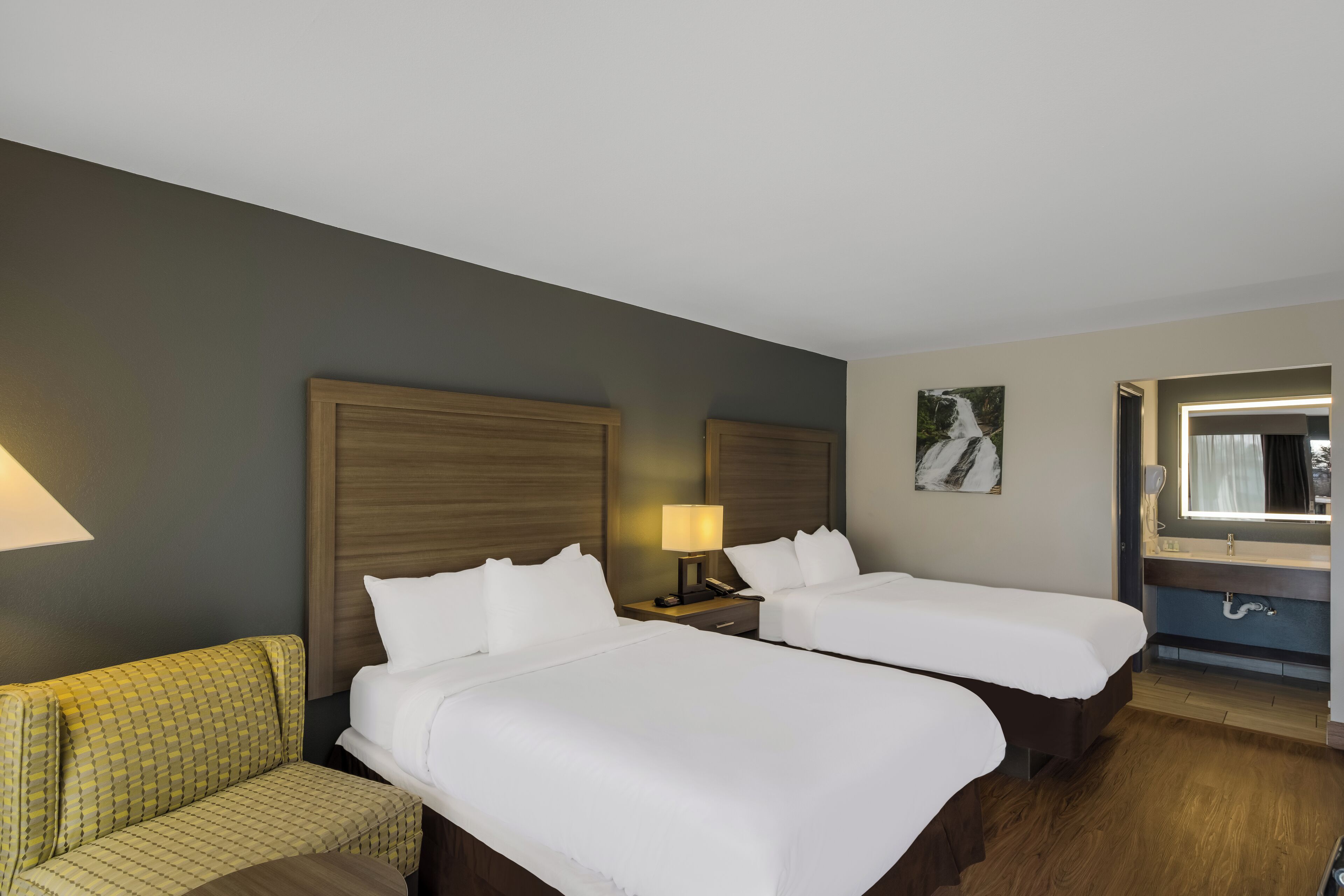 A hotel room with two beds, a sofa, a bedside table with a lamp, a mirror, and a painting on the wall.
