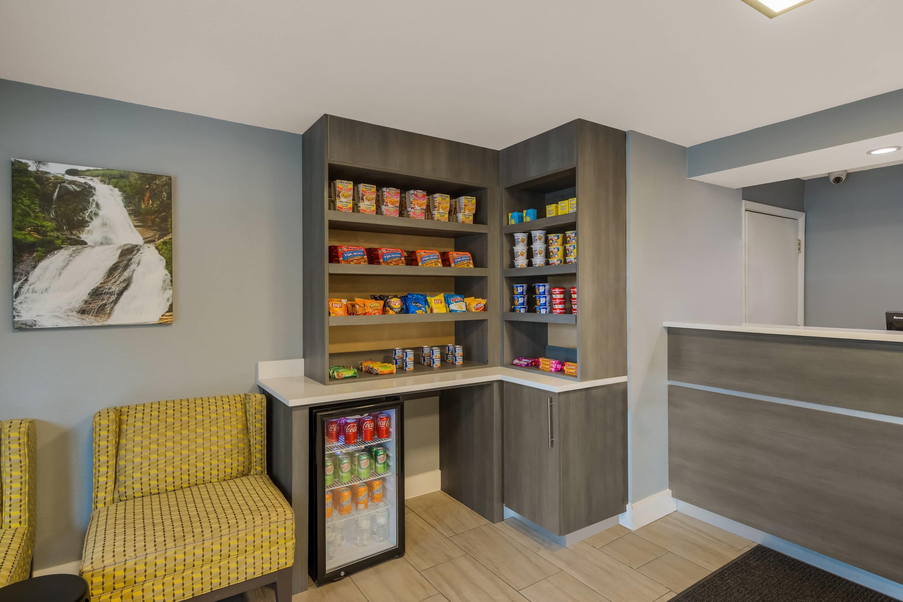 Una kitchenette con un refrigerador, estantes llenos de bocadillos y una zona de estar con una silla amarilla estampada.