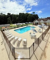 Un área de piscina con sillones de descanso y sombrillas, rodeada por una valla, con un edificio al fondo.