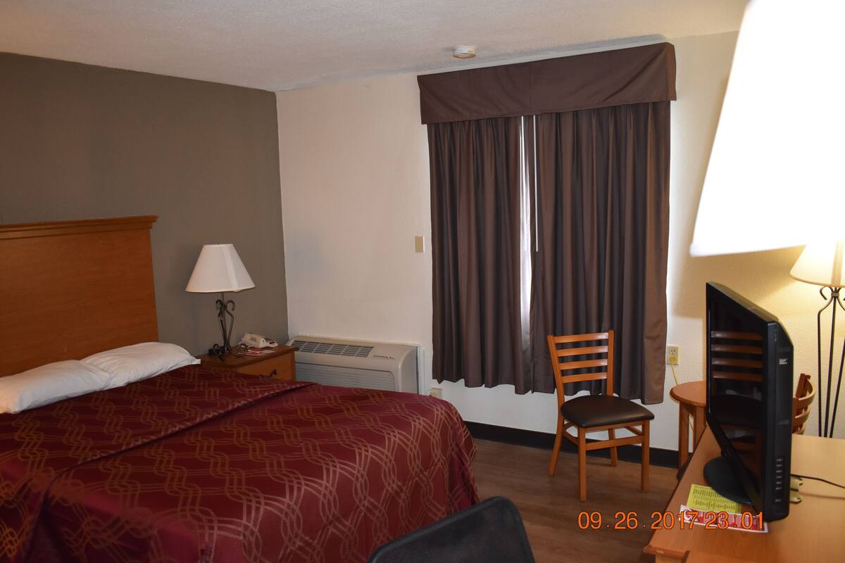Une chambre d’hôtel avec un lit, un bureau avec un ordinateur, une chaise, une lampe et une fenêtre avec des rideaux.