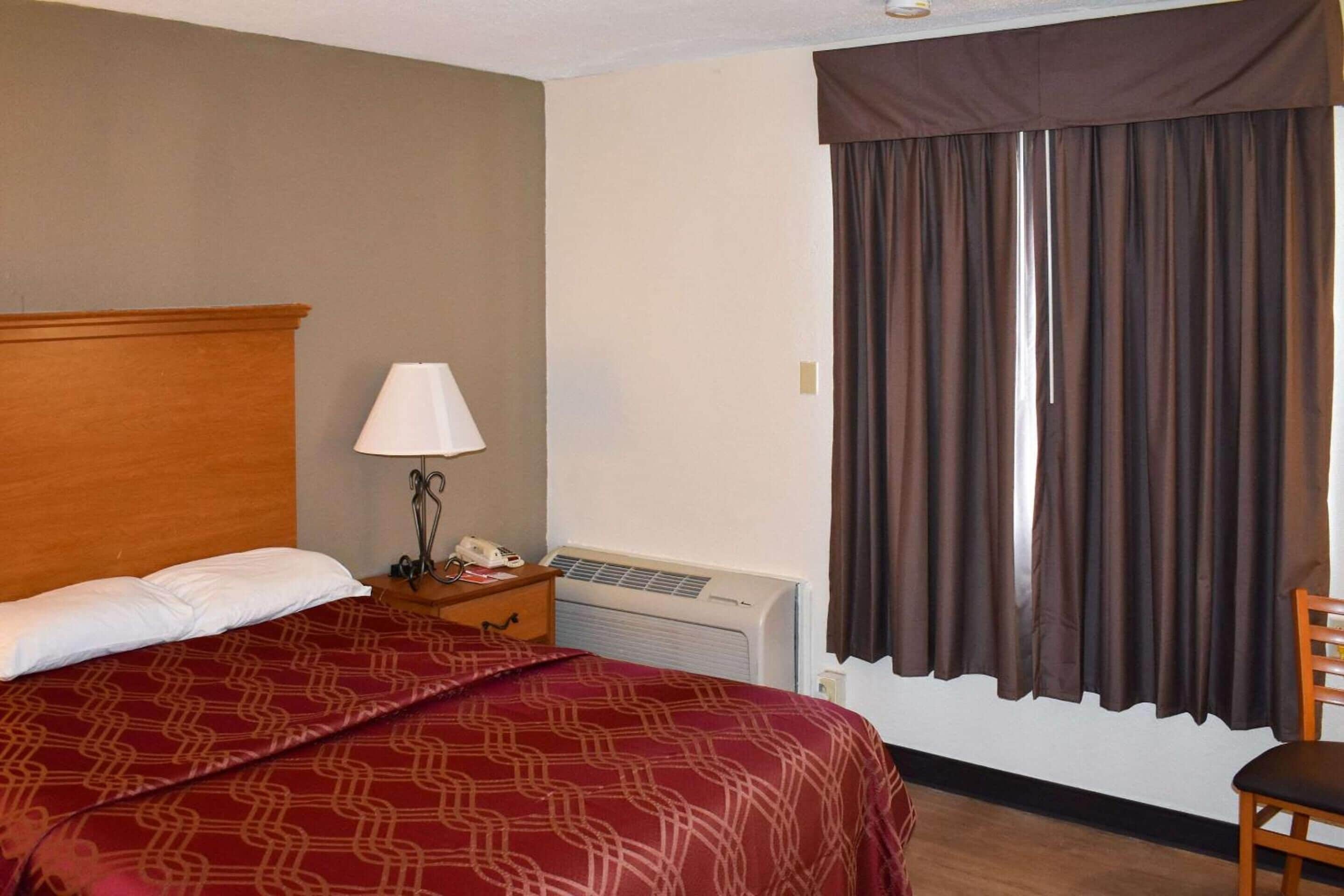 Habitación de hotel con cabecera de madera, cama con colcha roja estampada, mesita de noche con lámpara y teléfono, unidad de aire acondicionado y ventana con cortinas marrones.