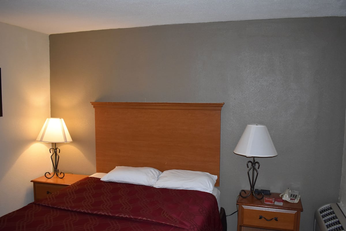 Une chambre d’hôtel avec une tête de lit en bois, un lit avec une literie blanche et rouge, deux tables de chevet avec des lampes, un petit tiroir et un téléphone fixé au mur.