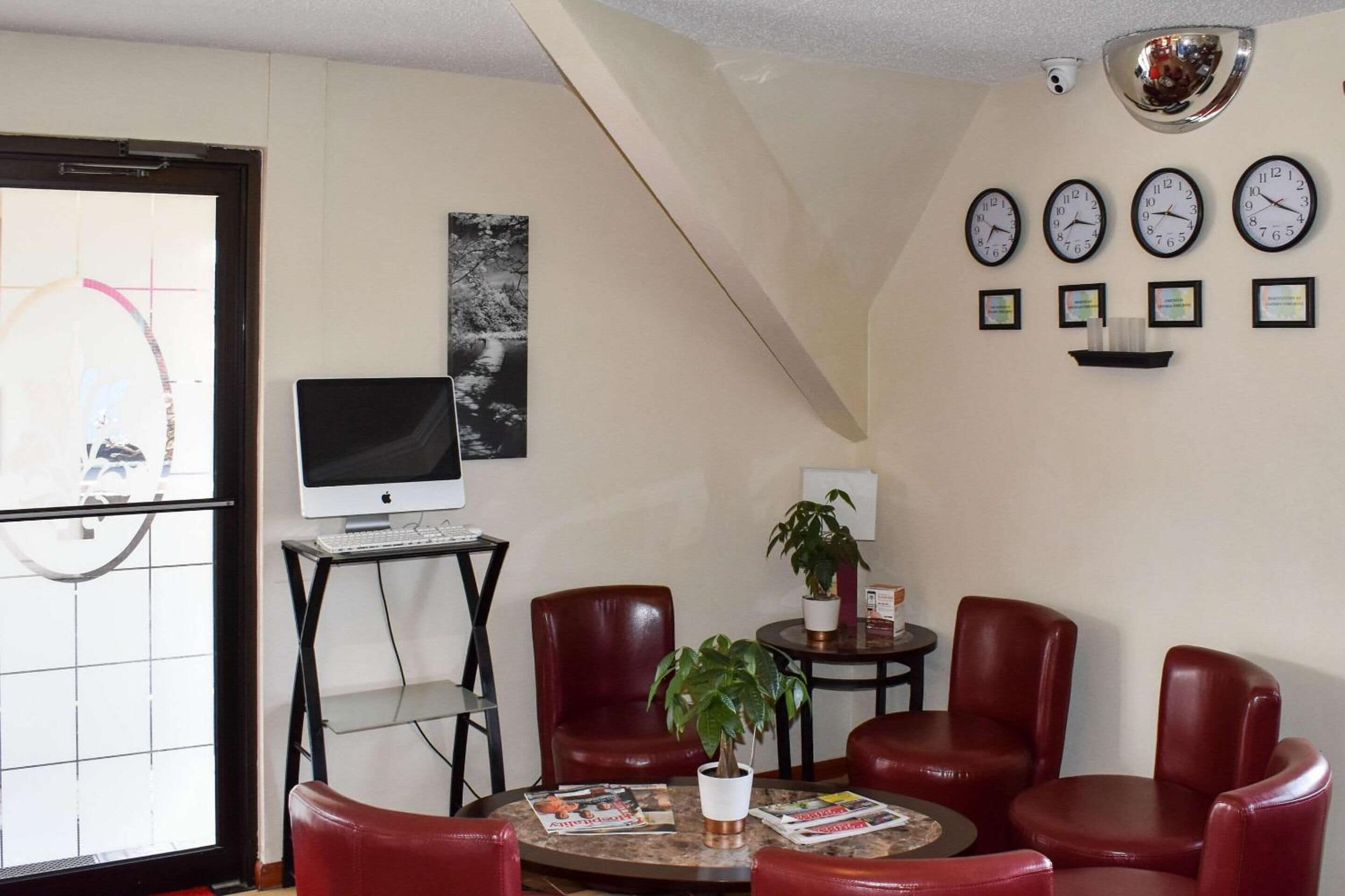A room with a desk, computer, red chairs, a small table with a plant, and four wall clocks.