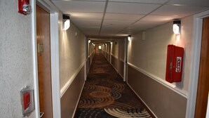 A hotel hallway with carpeted floors, white walls, and a red fire extinguisher on the right. - MHO Hotel Bordentown (Bordentown)